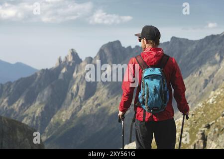 Ein bärtiger Mann mit Sonnenbrille und einer Mütze mit Rucksack steht auf einem Felsen und blickt in ein felsiges Tal hoch in den Bergen. Das Konzept der Tour Stockfoto