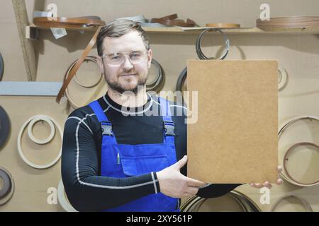 Der Hersteller von Möbeln in Spezialkleidung und Schutzbrille hält am Arbeitsplatz eine leere Platte für den Text. Handwerkliche Herstellung von Möbeln Stockfoto