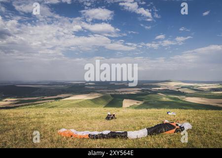 Ein professioneller Gleitschirm in voller Ausrüstung und ein Helm liegt und ruht auf dem Gras hoch in den Bergen und schaut auf die Wolken Stockfoto