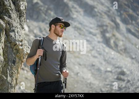 Der Träger eines stilvollen Hipster-Reisenden mit Bart und Rucksack in Sonnenbrille und einer Mütze mit Trekkingstöcken steht auf einem Felsen vor der Kulisse Stockfoto