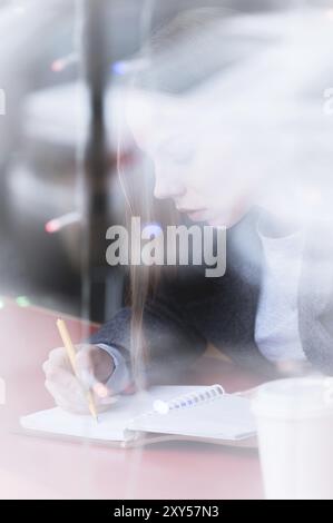 Porträt einer attraktiven jungen Journalistin mit Stift und Notizblock im Café hinter dem Schaufenster. Kontrastfreie Sicht durch Glasreflexion Stockfoto