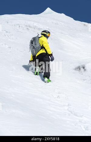 Ein Skifahrer in Helm und Maske mit Rucksack steigt auf einer Piste vor dem Hintergrund von Schnee und einem Gletscher. Backcountry Freeride Stockfoto