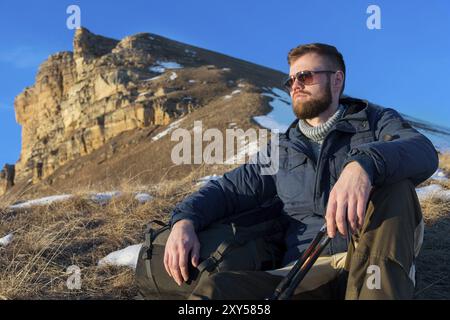 Porträt des ruhigen Hipster-Reisenden mit Bart in der Sonnenbrille sitzt auf der Natur. Ein Mann wandert in den Bergen mit Rucksack und skandinavischem Wa Stockfoto