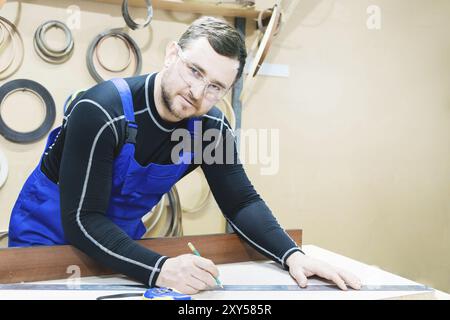 Ein bärtiger, müder, gutaussehender Zimmermann auf einem Tisch mit Bleistift zeichnet ein Schild auf das Brett. Am Arbeitsplatz gibt es einen gewöhnlichen Meister in blau gekleidet ca. Stockfoto