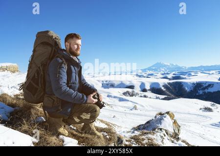 Porträt Ein bärtiger Hipster-Fotograf mit Rucksack und Sonnenbrille sitzt mit einer DSLR-Kamera in den Händen vor dem Hintergrund des Schnees Stockfoto