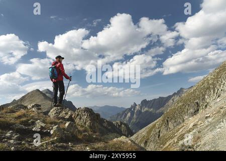 Ein bärtiger Mann mit Sonnenbrille und einer Mütze mit Rucksack steht auf einem Felsen und blickt in ein felsiges Tal hoch in den Bergen. Das Konzept der Tour Stockfoto