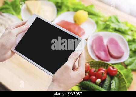 Nahaufnahme von oben auf weibliche Hände, die einen Tablet-Computer mit einem leeren Display halten, umgeben von gesunden Lebensmitteln auf einem Wohnküchentisch. Einzufügendes Werkstück Stockfoto