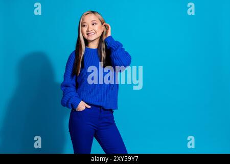 Porträt der jungen süßen chinesischen Frau im blauen gestrickten Pullover, der ihr gefärbtes schönes Haar tagträumt isoliert auf blauem farbigem Hintergrund berührt Stockfoto