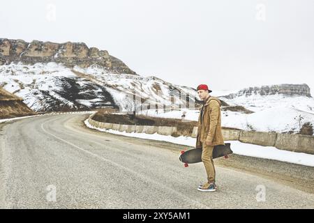 Stilvoller Happy Young man in einer Mütze und Hose, die auf einer Bergstraße stehen, ein Longboard in den Händen, das Leben genießen Stockfoto