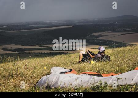 Ein professioneller Gleitschirm in voller Ausrüstung und ein Helm liegt und ruht auf dem Gras hoch in den Bergen und schaut auf die Wolken Stockfoto