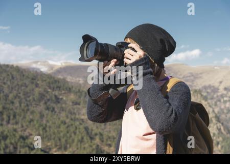 Nahaufnahme eines blonden Mädchens mit einer digitalen Spiegelreflexkamera, die in den Bergen fotografiert. Stylischer freiberuflicher Fotograf mit Rucksack in den Bergen Stockfoto