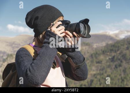 Nahaufnahme eines blonden Mädchens mit einer digitalen Spiegelreflexkamera, die in den Bergen fotografiert. Stylischer freiberuflicher Fotograf mit Rucksack in den Bergen Stockfoto