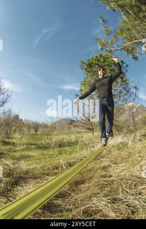 Ein Mann, mit Bart und Sonnenbrille alt, balanciert auf einer Slackline zwischen zwei Bäumen bei Sonnenuntergang am blauen Himmel Stockfoto