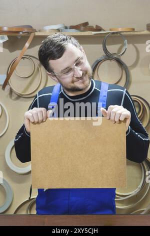 Der Hersteller von Möbeln in Spezialkleidung und Schutzbrille hält am Arbeitsplatz eine leere Platte für den Text. Handwerkliche Herstellung von Möbeln Stockfoto