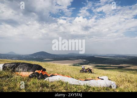 Ein professioneller Gleitschirm in voller Ausrüstung und ein Helm liegt und ruht auf dem Gras hoch in den Bergen und schaut auf die Wolken Stockfoto