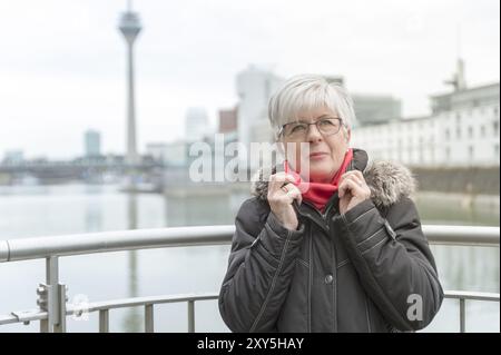 Eine Seniorin mit kurzen weißen Haaren steht im Medienhafen mit dem Rücken zum Rheinturm und betrachtet die umliegende Architektur Stockfoto