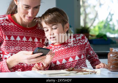 Mutter und Sohn schauen sich ein Rezept für Weihnachtssüße mit Schokolade auf dem Handy an, während sie in der Küche zuhause ein Dessert zubereiten Stockfoto