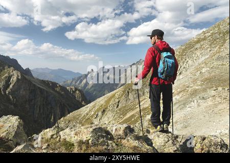 Ein bärtiger Mann mit Sonnenbrille und einer Mütze mit Rucksack steht auf einem Felsen und blickt in ein felsiges Tal hoch in den Bergen. Das Konzept der Tour Stockfoto