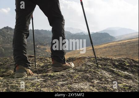 Hipster in Mütze und roter Jacke mit Stöcken für Nordic Walking mit Rucksack und gefaltetem Teppich zum Entspannen steht in den Bergen gegen das B Stockfoto