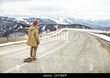 Stylischer Happy Young man in einer Mütze und Hose Jogginghose, die auf einem Longboard eine Bergstraße hinunterrollt und das Leben genießt Stockfoto