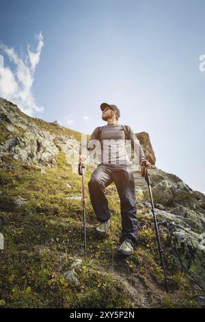 Ein bärtiger Mann mit Sonnenbrille und einer Mütze mit Rucksack steht auf einem Felsen und blickt in ein felsiges Tal hoch in den Bergen. Das Konzept der Tour Stockfoto