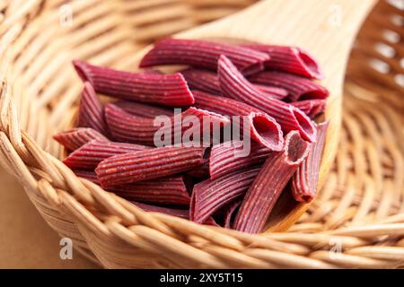 Rote Rüben-Penne-Pasta. Rohe glutenfreie Zutat. Diät ungekochte Makkaroni in rustikaler Rattanschale mit Holzlöffel. Stockfoto