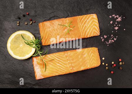 Roher Lachs mit Gewürzen auf schwarzem Hintergrund. Zwei Scheiben frischer Fisch, Schuß von der Oberseite mit Zitrone, Rosmarin, Salz und Pfeffer Stockfoto