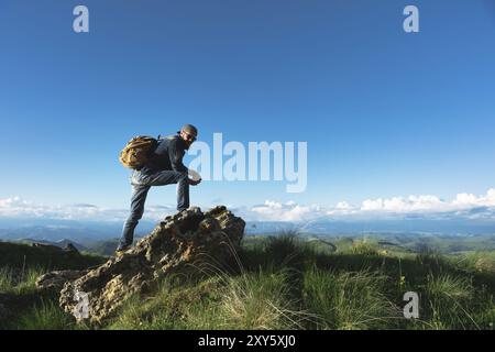 Stilvoller bärtiger Reisender in Sonnenbrille und einer Mütze mit Rucksack in Denim-Anzug und gelben Schuhen steht auf einem großen Stein vor dem Hintergrund Stockfoto