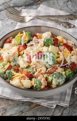 Gedämpfter Brokkoli, Blumenkohlsalat mit Speck, Zwiebeln, Käse und Mayonnaise-Dressing aus der Nähe auf dem Teller auf dem Tisch. Vertikal Stockfoto