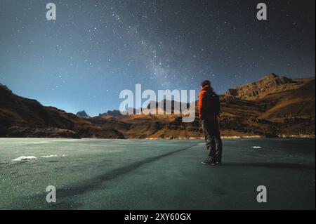 Ein brütender Mann steht auf der Oberfläche eines gefrorenen Sees, beleuchtet von Mondlicht hoch in den Bergen vor der Kulisse epischer Felsen. Das Konzept o Stockfoto