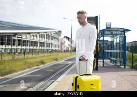 Gutaussehender kaukasischer Geschäftsreisender mit Tasche und Koffer sitzen an der Bushaltestelle des öffentlichen Nahverkehrs und warten mit Handy Stockfoto