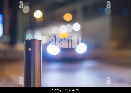 Blick über einen Poller am Straßenrand auf ein fahrendes Auto mit eingeschalteten Scheinwerfern bei Nacht Stockfoto