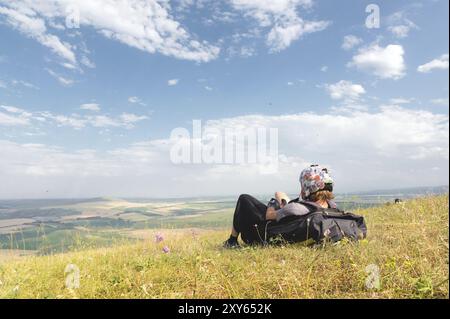 Ein professioneller Gleitschirm in voller Ausrüstung und ein Helm liegt und ruht auf dem Gras hoch in den Bergen und schaut auf die Wolken Stockfoto