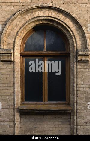 Vintage Bogenfenster in der Wand aus gelbem Ziegelstein. Schwarzes Glas in einem dunkelroten Holzrahmen. Das Konzept der antiken Vintage-Architektur im Bau Stockfoto