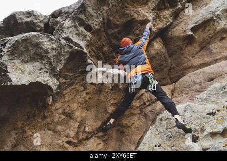 Hipster, Kletterer im Alter, in dem man einen wunderschönen Felsen ohne Versicherung und Helm hochklettert. Ein Bergsteiger in Hut und Daunenjacke mit Tasche für magnes Stockfoto