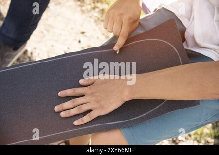 Ein kleiner Junge auf den Knien klebt das Griptape in Begleitung von Freunden bei sonnigem Wetter auf ein Skateboard. Vorbereitung eines Skateboards für einen Skatepark-Wettkampf Stockfoto