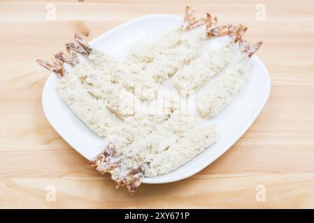 Nahaufnahme von rohen Tempura-Garnelen in panierter Form lag auf einem weißen Teller, der auf einem Holztisch steht. Halbfertige Meeresprodukte für die Hausmannskost Stockfoto