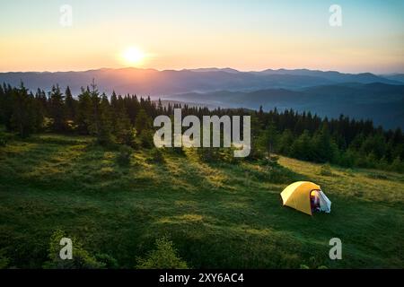 Blick aus der Vogelperspektive auf das gelbe Zelt auf einem grasbewachsenen Hügel, beleuchtet von aufgehender Sonne. Atemberaubendes Panorama von bewaldeten Bergen und sanften Hügeln unter klarem Himmel, die einen ruhigen und malerischen Campingplatz schaffen. Stockfoto