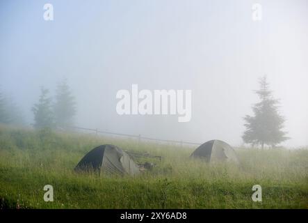 Zwei Zelte auf einem grasbewachsenen Hügel, umgeben von dickem Morgennebel. Die Umrisse von Kiefern und Holzzaun sind durch Nebel leicht sichtbar und schaffen eine ätherische, ruhige Atmosphäre. Stockfoto