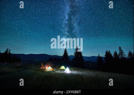 Verschwommene Silhouetten von Touristen, die auf dem Campingplatz am Lagerfeuer ruhen, mit leuchtenden Zelten in der Nähe unter Sternenhimmel. Die Milchstraße erstreckt sich über die Berge und wirft magisches Licht über die beschauliche Berglandschaft. Stockfoto