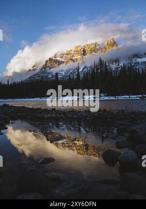 Ein ruhiger Fluss reflektiert schneebedeckte Berge und Kiefernwälder unter einem bewölkten, bei Sonnenuntergang beleuchteten Himmel, umgeben von Wolken, die sich im Fluss, Castle Mountain, Banff, spiegeln Stockfoto