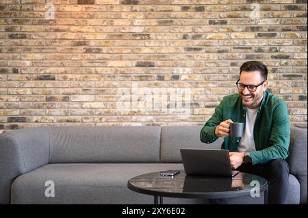 Positiver Geschäftsmann, der im Büro auf dem Sofa sitzt und Kaffee trinkt und E-Mails über Laptop liest Stockfoto