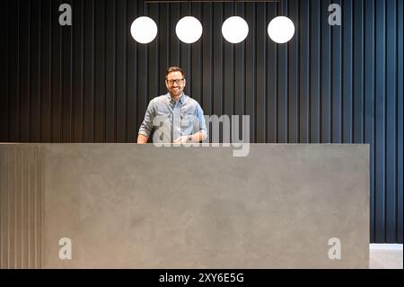 Selbstbewusster männlicher Rezeptionist mit Brille und Blick auf die Kamera, während er über der Wand am Lobby-Schreibtisch stand Stockfoto