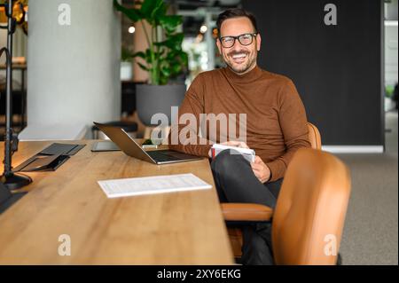 Porträt eines inspirierten männlichen Unternehmers in Brille, der über einem Laptop arbeitet, während er im Büro am Schreibtisch sitzt Stockfoto