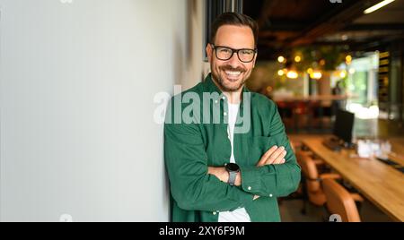 Porträt eines selbstbewussten Geschäftsmannes mit Brille und überkreuzten Armen, der sich im Büro an die weiße Wand lehnt Stockfoto