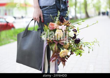 Junge, schöne Frau, die auf der Straße läuft mit Tasche und Blumenstrauß. Model mit stilvoller Kleidung und Accessoires Stockfoto