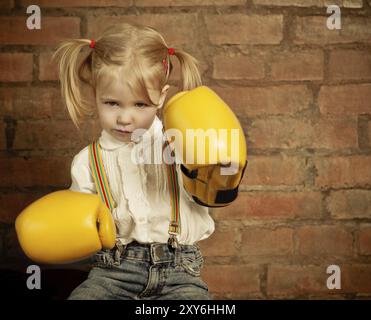 Kleines Mädchen mit gelben Boxhandschuhe über Ziegel Wand Hintergrund. Mädchen-Power-Konzept Stockfoto