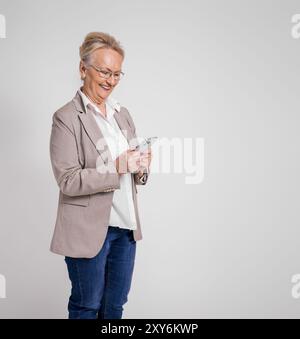 Lächelnde reife Unternehmerin, die Nachrichten über Mobiltelefon auf isoliertem weißem Hintergrund überprüft Stockfoto