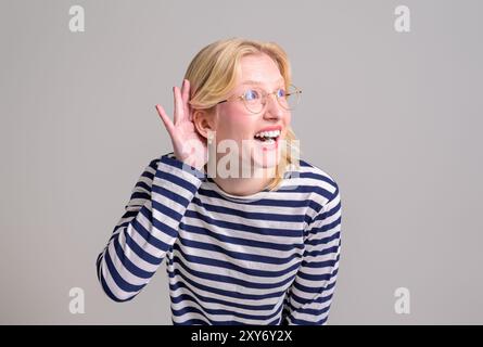 Neugierige glückliche, blonde junge Frau mit Hand nahe dem Ohr, die versucht, Klatsch über weißem Hintergrund zu hören Stockfoto