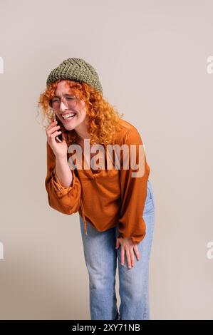 Glückliche junge Frau mit rothaarigem und gestricktem Hut, die fröhlich über Smartphone auf weißem Hintergrund spricht Stockfoto